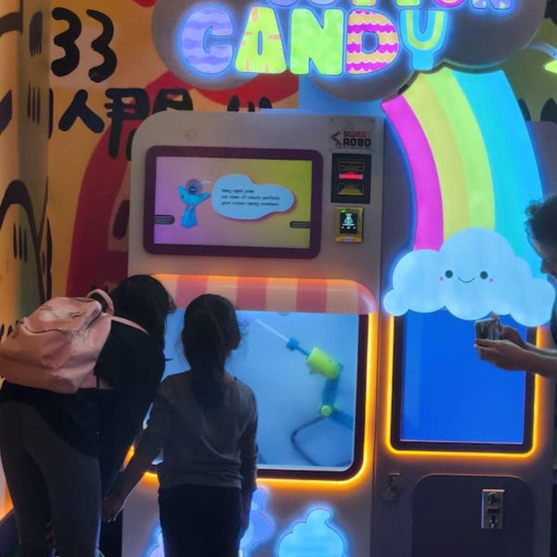 Two young ladies watch their cotton candy being made by the Sweet Robo Model VX Cotton Candy Vending Machine