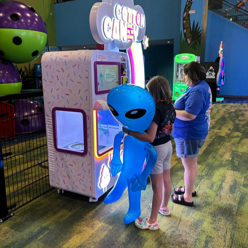A mother and her child are waiting for their cotton candy to be made from a Sweet Robo Model VX Cotton Candy Vending Machine, while at an arcade